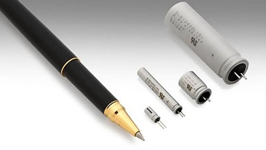 A sleek black fountain pen with gold accents is displayed alongside various capacitors of different sizes. The capacitors feature visible text and numeric values, including '47 µF' and '25 V'. 