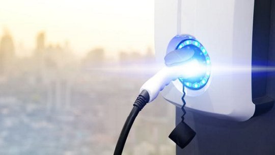 Une vue rapprochée d'une station de recharge pour véhicules électriques avec une lumière bleue brillante indiquant que la recharge est en cours. Le câble de recharge est solidement connecté à la station. L'arrière-plan présente une silhouette urbaine floue, suggérant un environnement urbain. L'image met en avant l'énergie propre et la technologie moderne.