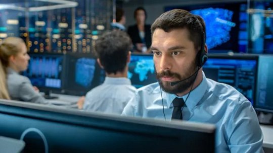 A group of professionals are seated at computer workstations in a high-tech security operations center. Multiple monitors display data and digital maps, suggesting a focus on cybersecurity or network monitoring.