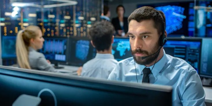 A group of professionals are seated at computer workstations in a high-tech security operations center. Multiple monitors display data and digital maps, suggesting a focus on cybersecurity or network monitoring.