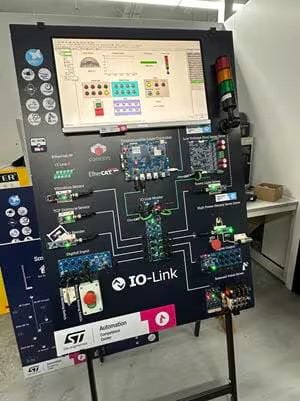 A large demonstration board showcases an IO-Link industrial automation system, featuring various electronic components and wiring. The board includes a digital display at the top showing a control interface, with multiple buttons, switches, and indicator lights visible.