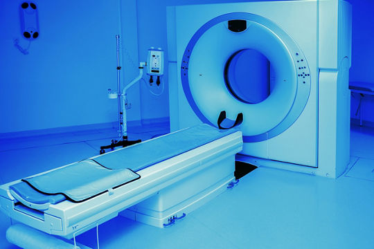 A contemporary CT scanner positioned in a medical examination room. The setting features a clean, sterile environment with blue lighting that emphasizes the equipment's design.