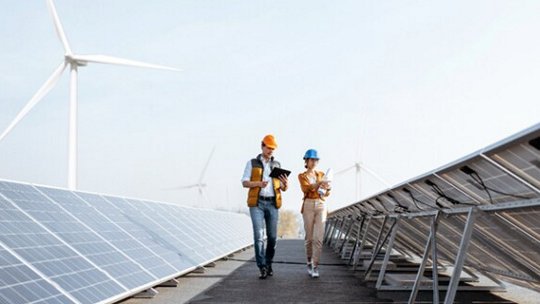 Two engineers wearing safety helmets and vests walk between rows of solar panels, conducting an inspection. The setting is an outdoor renewable energy facility with visible wind turbines in the background.