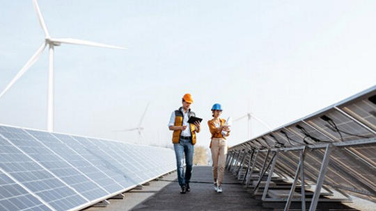 Two engineers wearing safety helmets and vests walk between rows of solar panels, conducting an inspection. The setting is an outdoor renewable energy facility with visible wind turbines in the background.