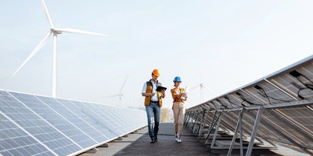 Two engineers wearing safety helmets and vests walk between rows of solar panels, conducting an inspection. The setting is an outdoor renewable energy facility with visible wind turbines in the background.