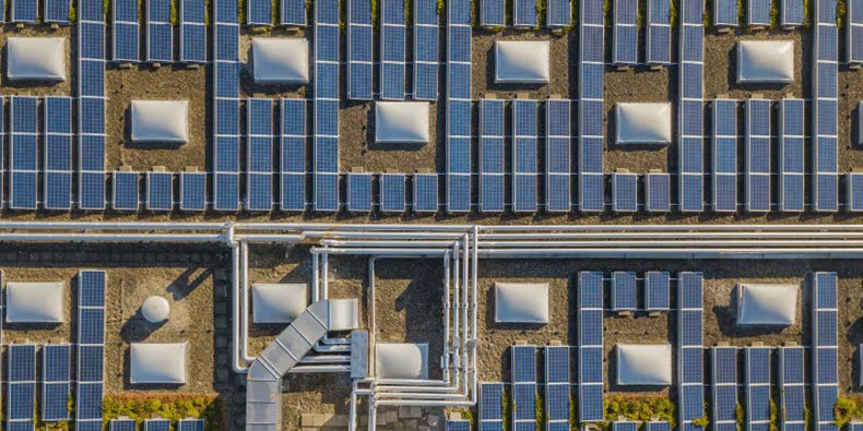 This image shows an array of solar panels installed on a flat rooftop, viewed from above. The panels are arranged in neat rows with visible ventilation units and metal piping running across the roof.