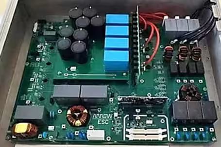 This image shows a close-up view of an electronic circuit board inside a metal enclosure. The board features several large capacitors, blue rectangular components, and multiple coils and relays. Red and black wires are connected to the upper right, and various connectors and chips are visible.