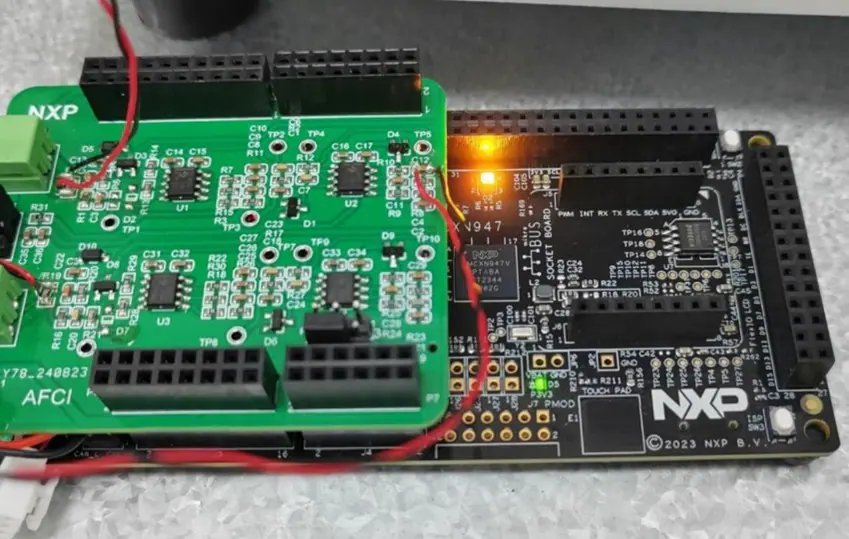 The image shows an NXP AFCI development board connected to a microcontroller base, with a green circuit board mounted on top. The board is powered on, as indicated by an illuminated orange LED.