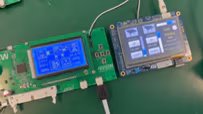 Two electronic circuit boards are shown side by side on a green surface, each featuring a digital display. The left board has a blue monochrome screen displaying technical diagrams and numbers, while the right board has a color touchscreen interface with multiple buttons and text. Both devices are connected by wires.
