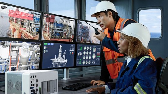 Two engineers wearing safety helmets and vests monitor multiple screens in a control room