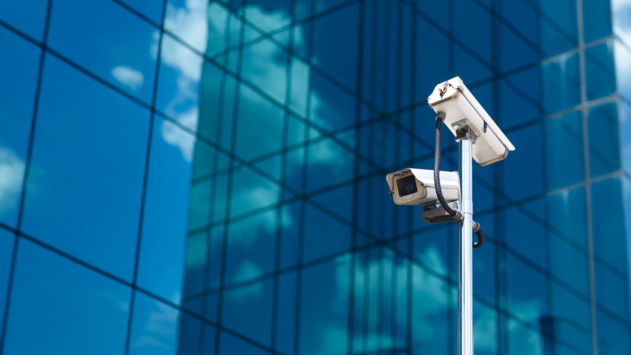 Two surveillance cameras are mounted on a tall metal pole in front of a modern glass office building.