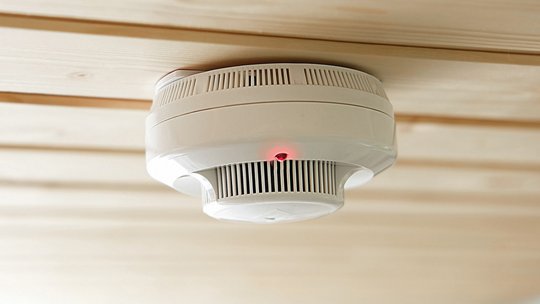 A white smoke detector is mounted on a light wooden ceiling. 