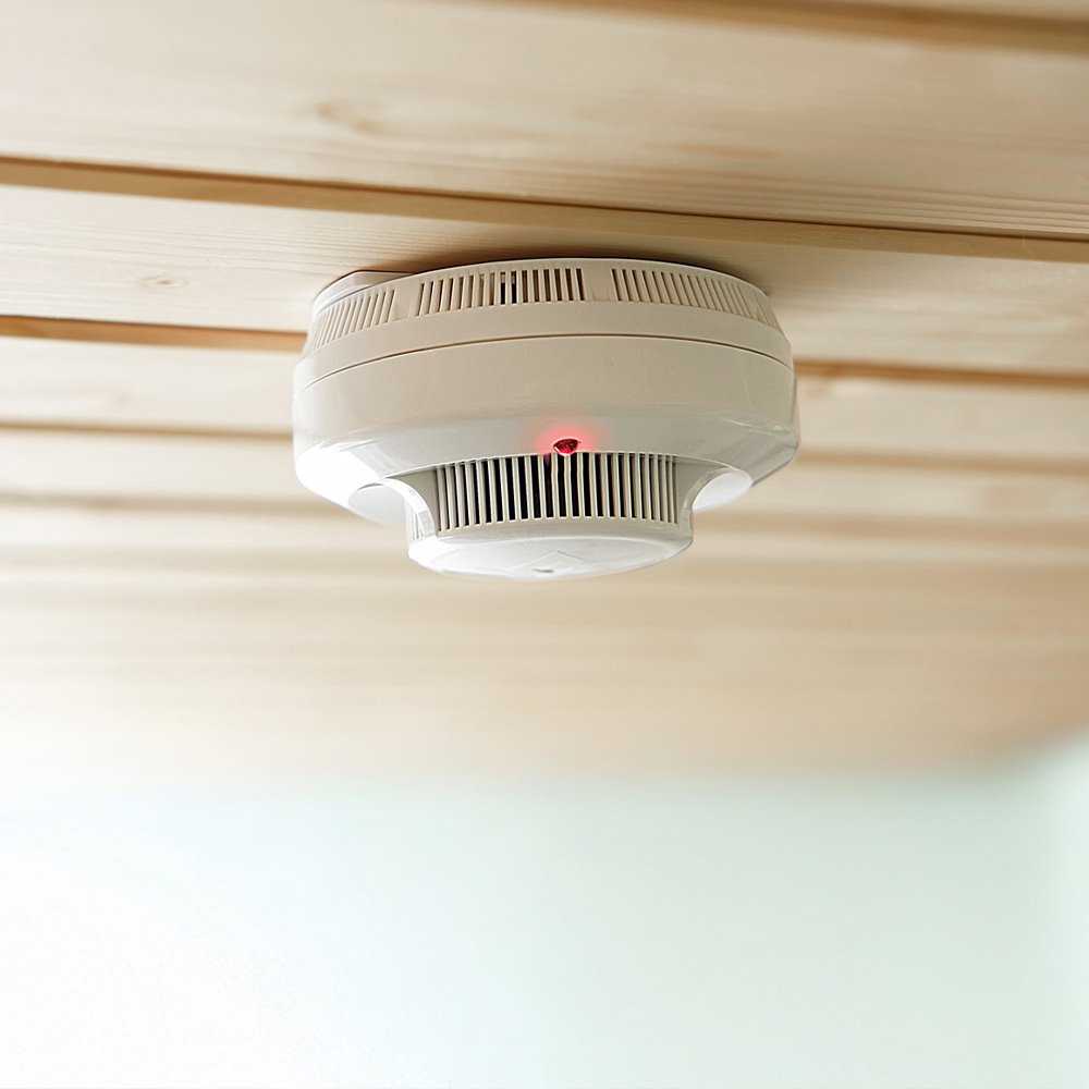 A white smoke detector is mounted on a light wooden ceiling. 