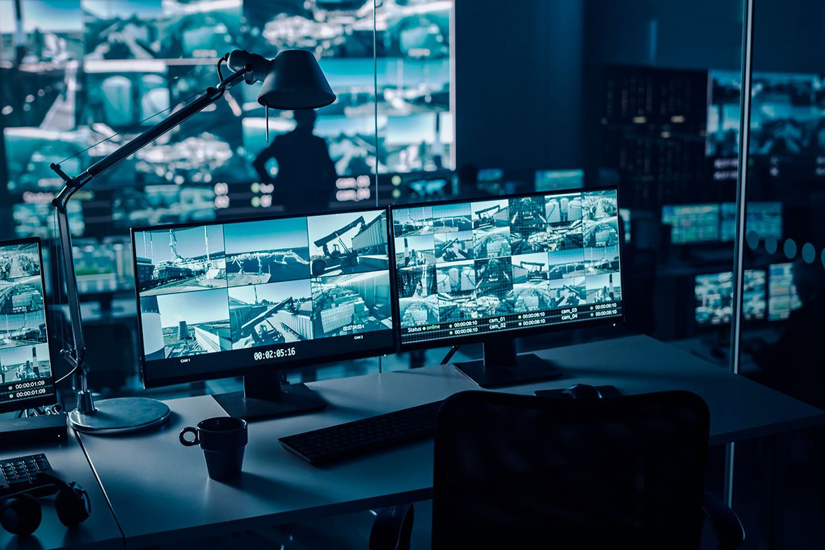 A modern security control room features multiple computer monitors displaying various CCTV camera feeds.