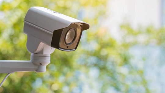 A white outdoor security camera is mounted on a metal arm, facing slightly downward. 