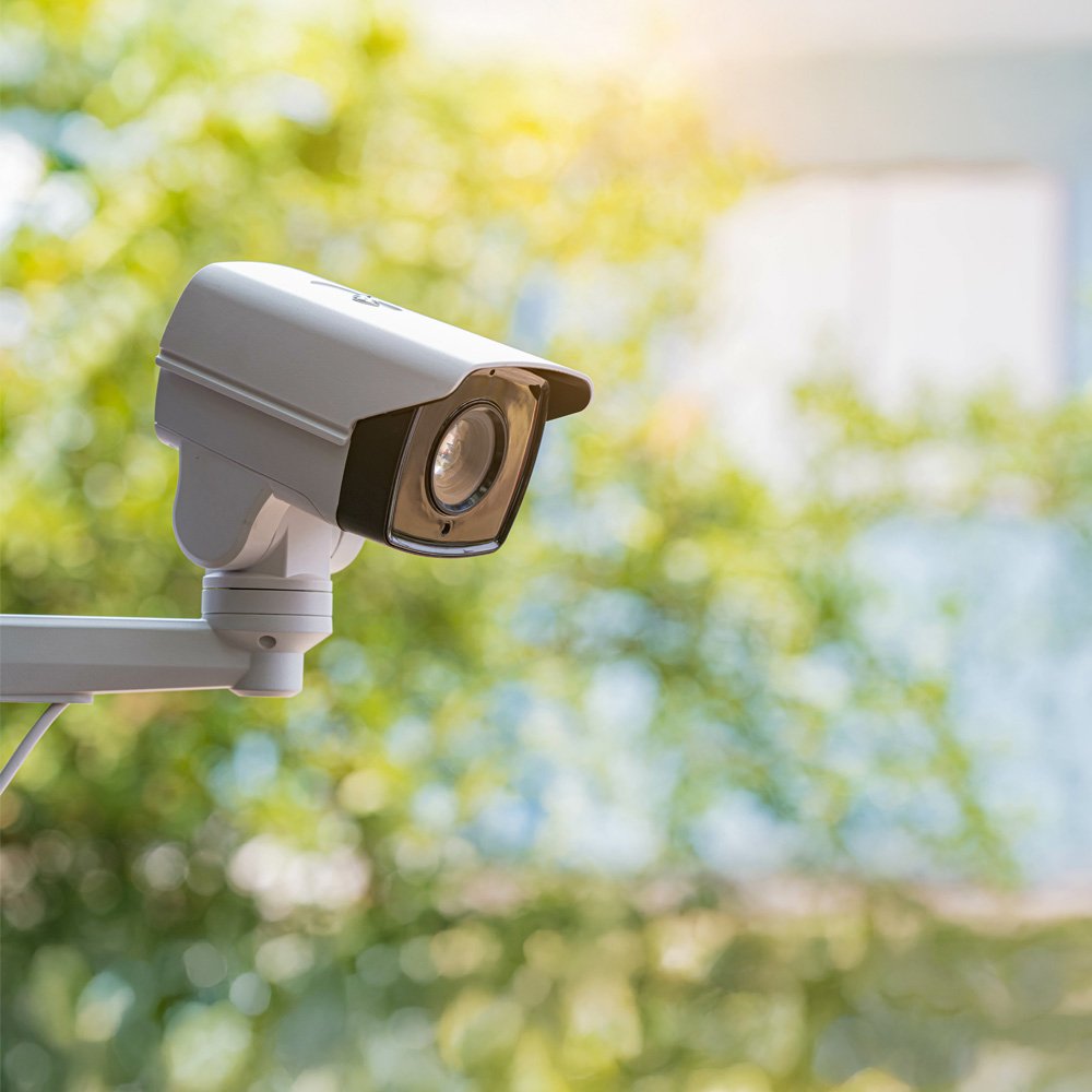 A white outdoor security camera is mounted on a metal arm, facing slightly downward. 