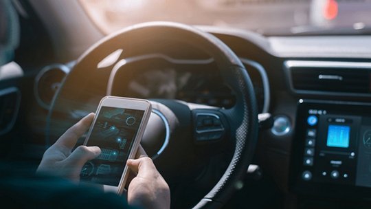 A person holds a smartphone while seated in a modern car interior. The dashboard displays illuminated digital screens with various controls and data.
