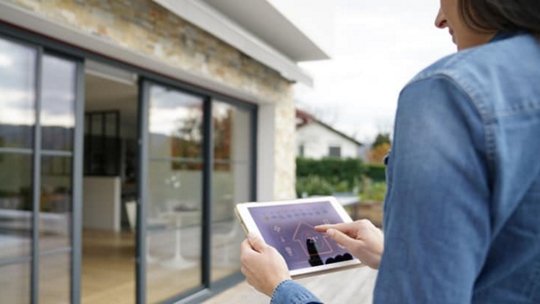 Una mujer interactúa con una tableta para gestionar las funciones de un hogar inteligente, de pie frente a una casa moderna con grandes puertas de vidrio.