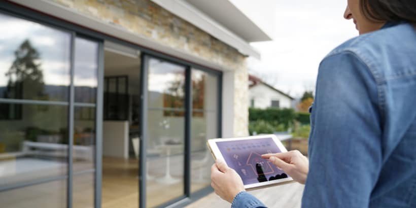 Una mujer interactúa con una tableta para gestionar características del hogar inteligente, de pie fuera de una casa moderna con grandes puertas de cristal.