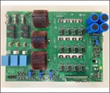 A green printed circuit board features multiple electronic components, including three large relays, several blue cylindrical capacitors, and various integrated circuits. The board is photographed from above, clearly displaying its layout and soldered connections.