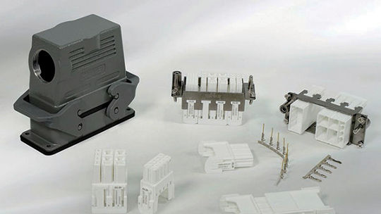 A collection of industrial connector components displayed on a neutral surface. The set includes a gray protective housing and multiple white connector modules with metallic pins. 