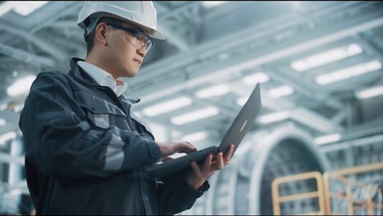 A professional wearing a white safety helmet and dark work jacket operates a laptop in a modern industrial setting.
