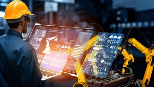 노란색 안전 헬멧을 착용한 남성 엔지니어가 산업 환경에서 로봇 팔을 제어하기 위해 디지털 인터페이스를 조작하고 있습니다.