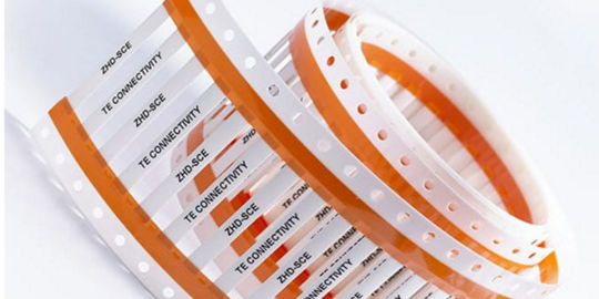 Rolls of orange and white TE Connectivity label tape with printed product information.