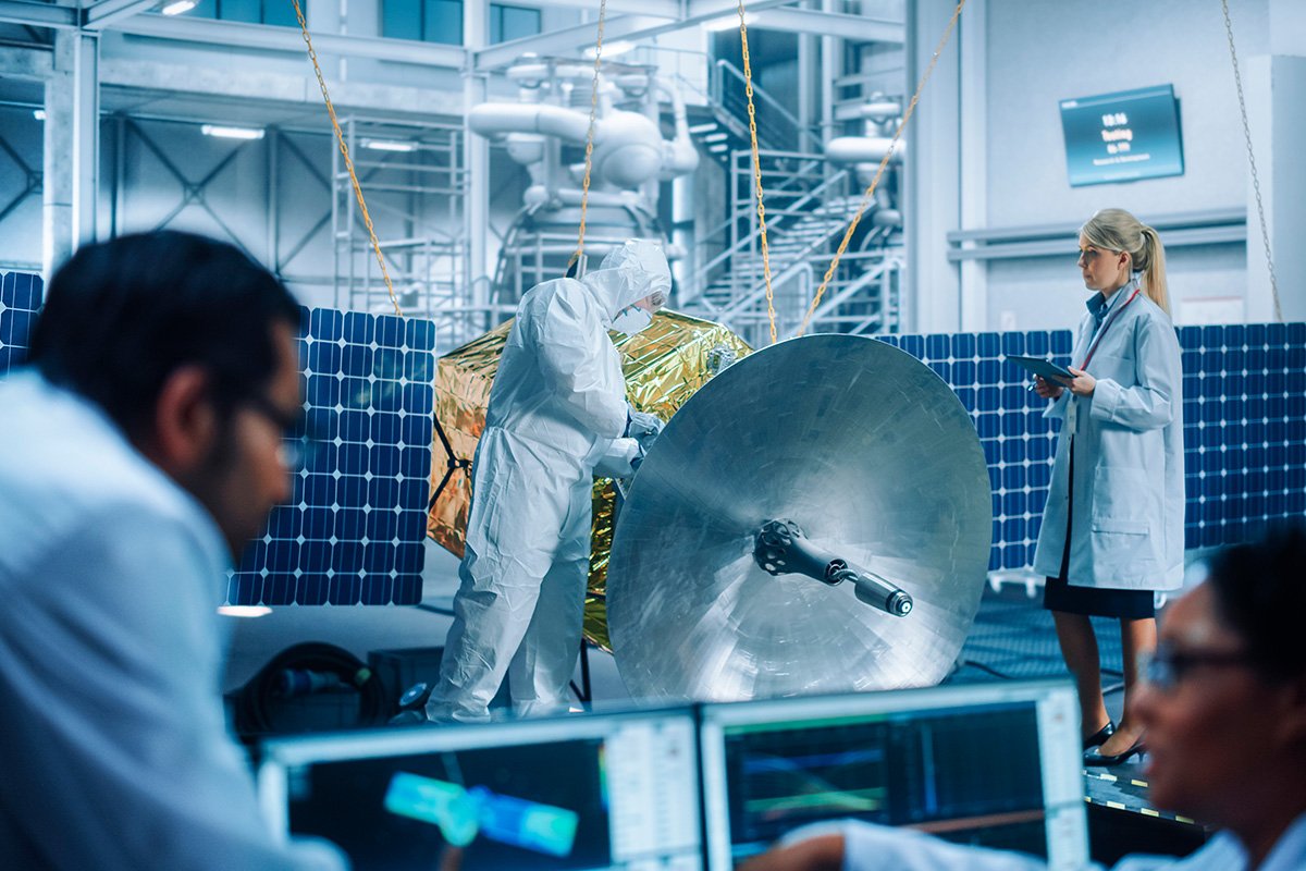 Scientists and engineers work together on a large satellite dish inside a high-tech laboratory. 