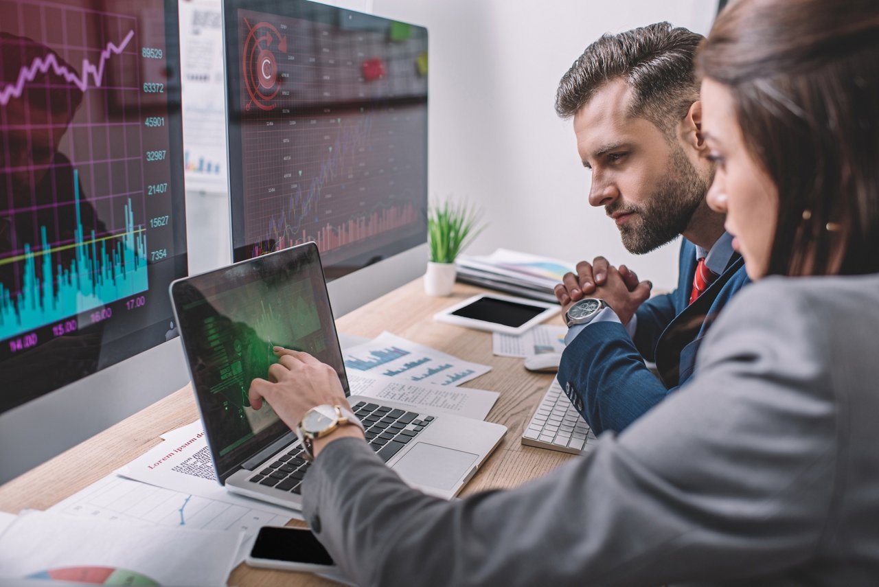 Two people working at a desk reviewing charts and graphs on a large monitor while discussing data displayed on a laptop.