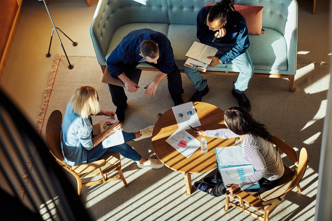 Overhead view of four people seated around a small round table in a modern office lounge, reviewing charts, reports, and notes during a collaborative meeting. Sunlight streams through windows, casting striped shadows across the room.