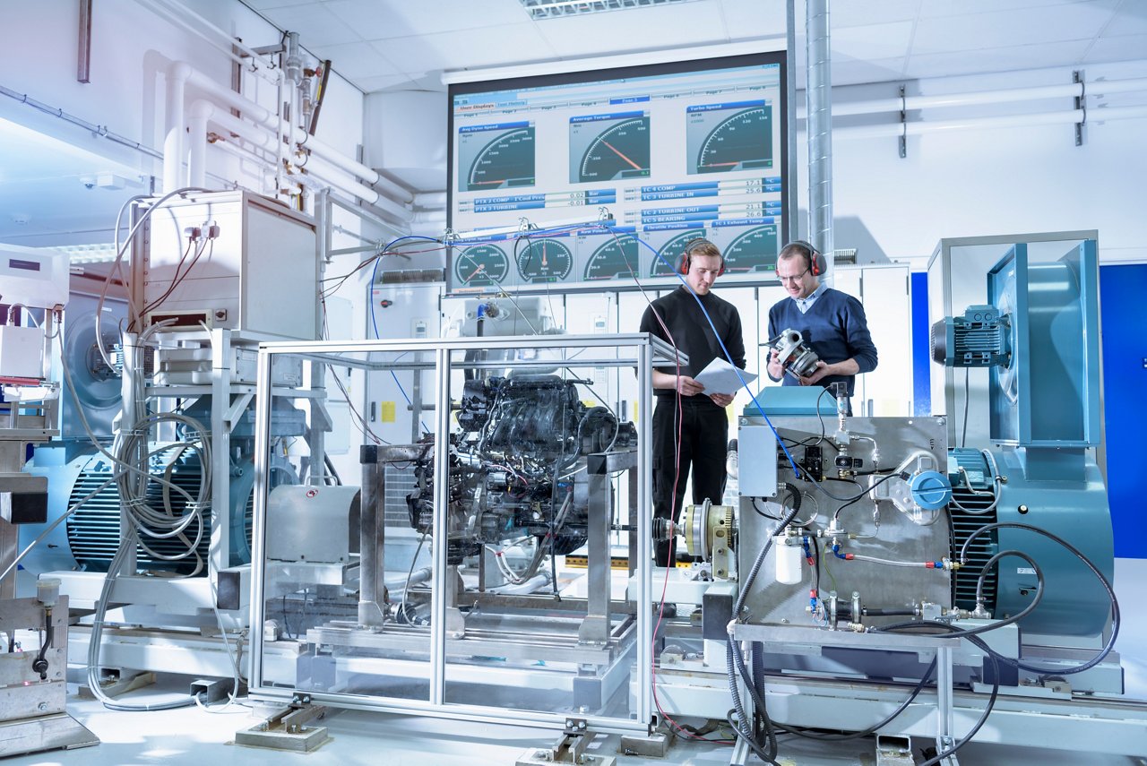 Two people standing in an industrial laboratory surrounded by large machinery, cables, and monitoring equipment, with data displays visible on a screen behind them.