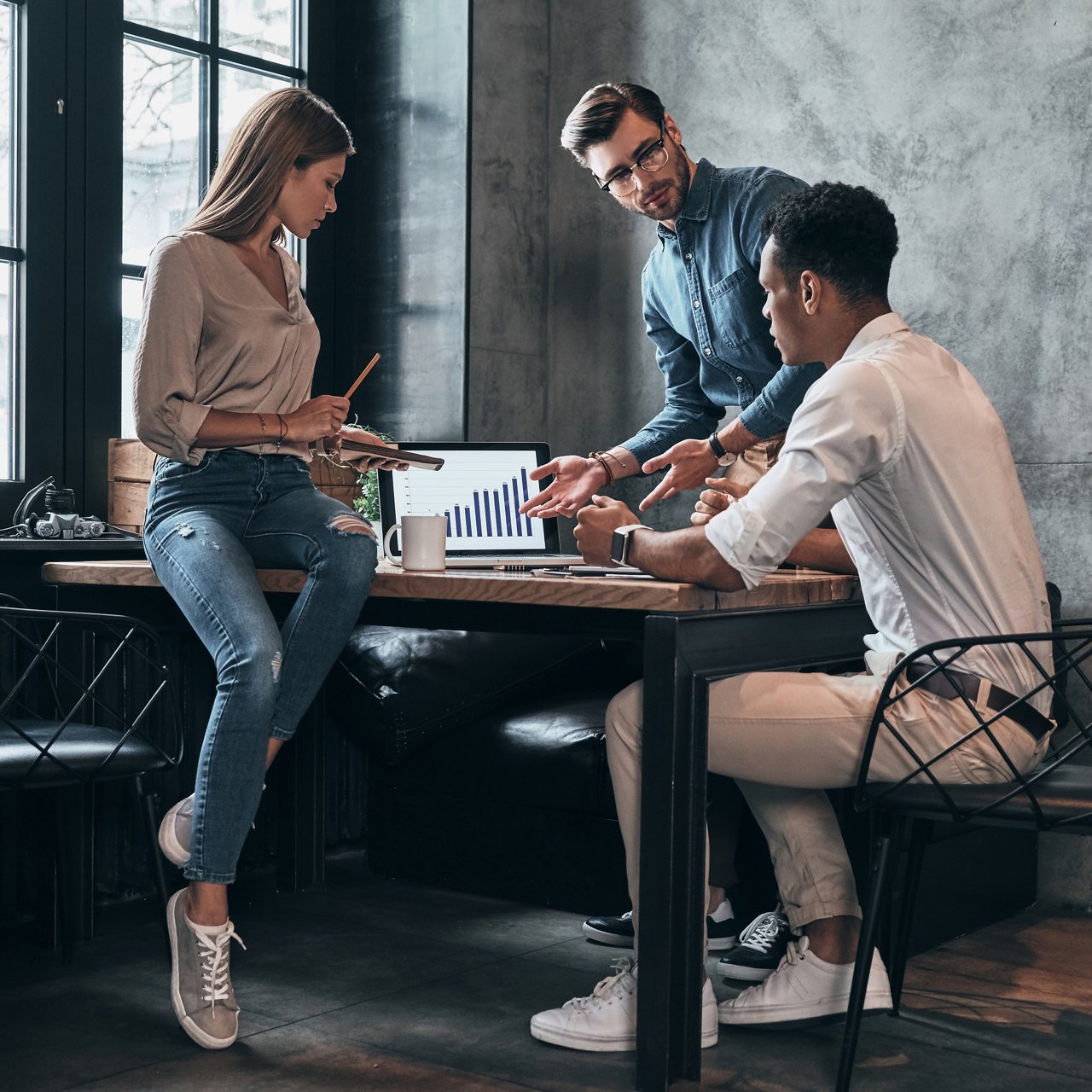 **Alt text (255 characters or less):**  Three colleagues meet around a table in a modern office, reviewing a laptop displaying an upward-trending bar chart. Notes, a tablet, and a coffee mug are nearby as the group discusses data and business performance.