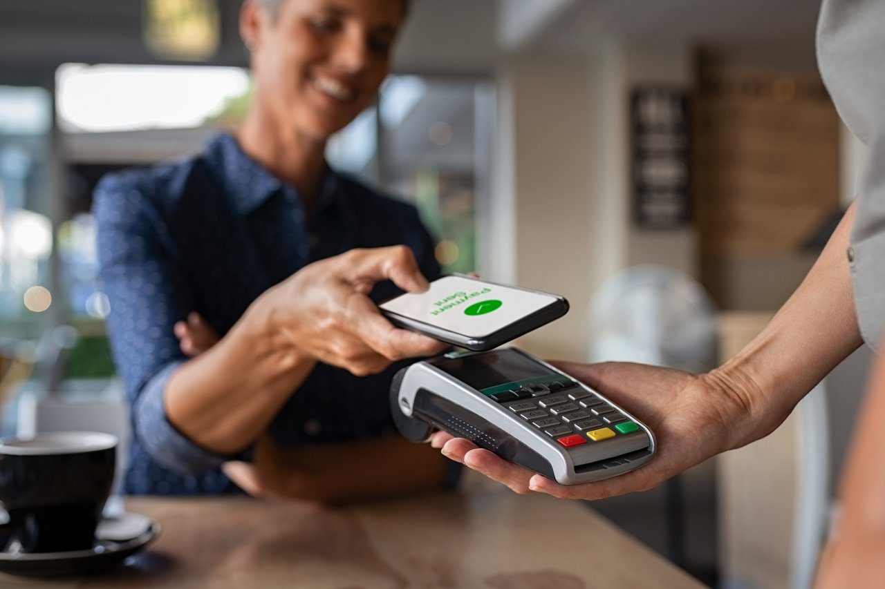 Person holds a smartphone over a contactless payment terminal to complete a digital transaction. The terminal is held across a table in a café or retail setting, illustrating mobile payment and tap-to-pay technology.