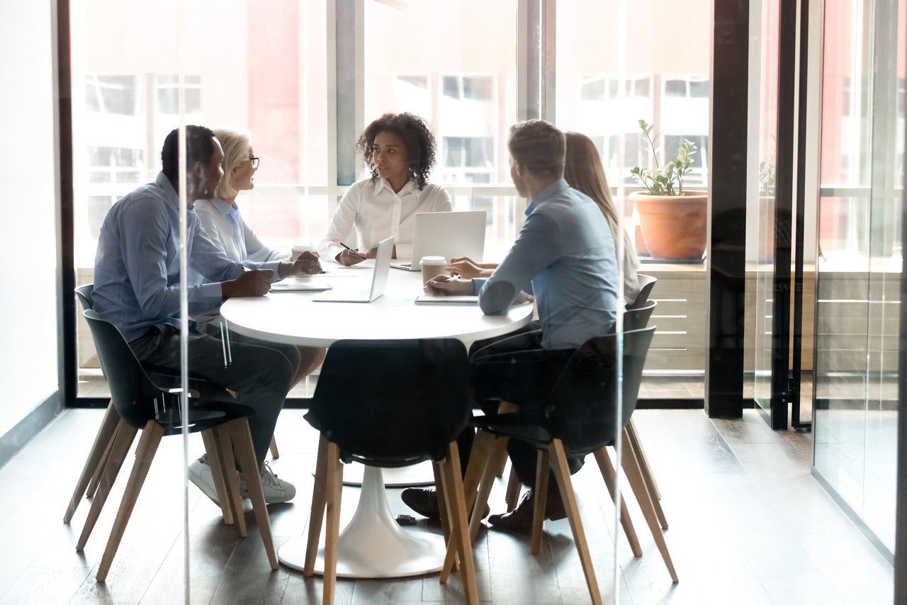 A group of people seated around a round table in a glass-walled meeting room, using laptops and having a discussion.