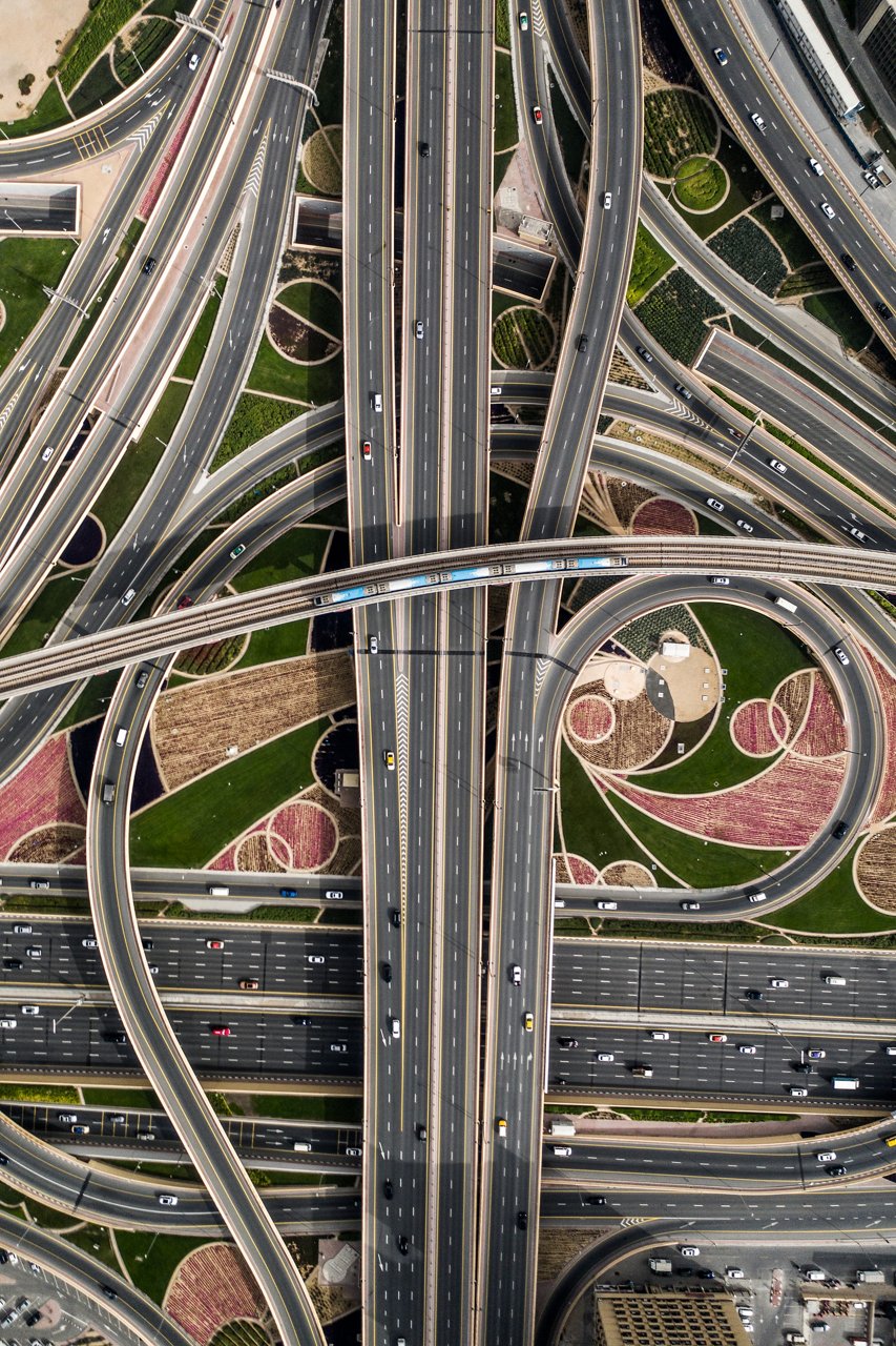 Aerial view of a large multi-level highway interchange with intersecting roads, looping ramps, and an elevated rail line crossing overhead. Vehicles travel along multiple lanes, surrounded by landscaped green spaces and geometric patterns.