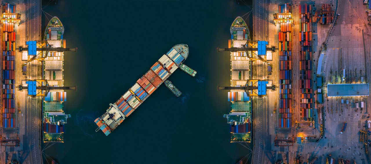 Aerial view of a container ship maneuvering between two illuminated cargo terminals at a busy port. Shipping containers, cranes, and dock infrastructure line both sides of the waterway, highlighting global freight and logistics operations.