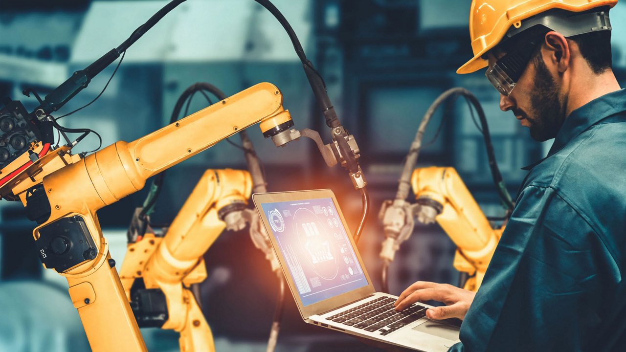 An industrial engineer uses a laptop to control and monitor robotic arms in a modern manufacturing facility. 