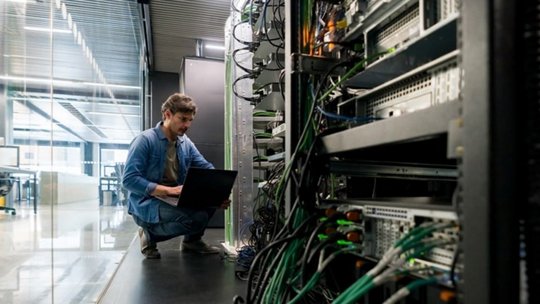 기술자가 현대적인 서버실에서 네트워크 장비 랙 사이에 있는 노트북을 작업하며 웅크리고 있습니다.