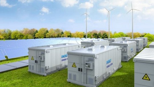 A row of energy storage containers is positioned in a green field surrounded by solar panels and wind turbines.