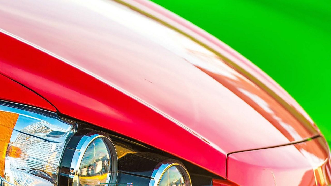 A vibrant close-up of a red car's front headlight, showcasing sleek design and reflective details. The background features a bright green setting, adding contrast to the image. 