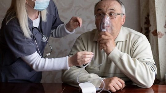 A healthcare professional wearing a mask is assisting a patient with a medical device in a home setting