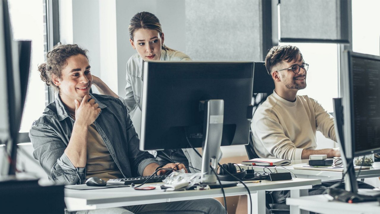 A group of professionals working together in a contemporary office environment. The scene features desks with computers, monitors, and office supplies, suggesting a focus on teamwork and productivity. 