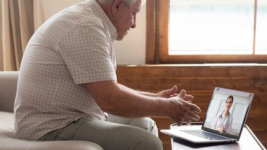 An elderly man sits in a cozy living room setting, engaging in a virtual consultation with a doctor displayed on a laptop screen
