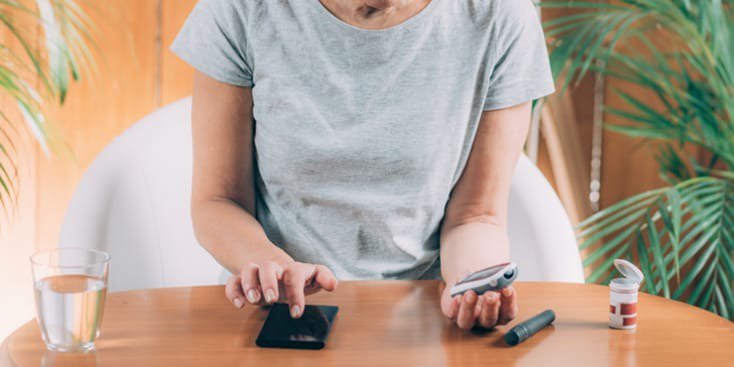 Una mujer de mediana edad está sentada en una mesa de madera, usando un dispositivo de monitoreo de glucosa y un teléfono inteligente.