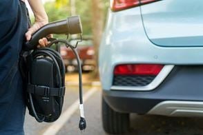 A close-up of a person holding an electric vehicle charging cable near a parked light blue car. 