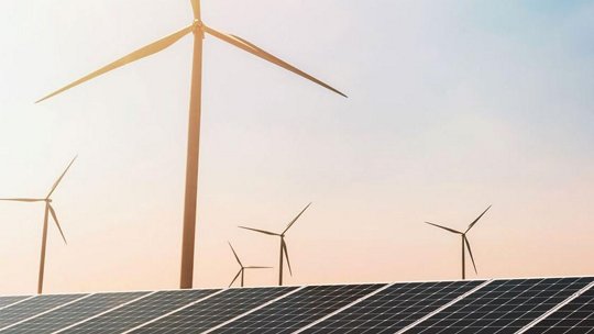 Una instalación de energía renovable con filas de paneles solares en primer plano y múltiples turbinas eólicas en el fondo.