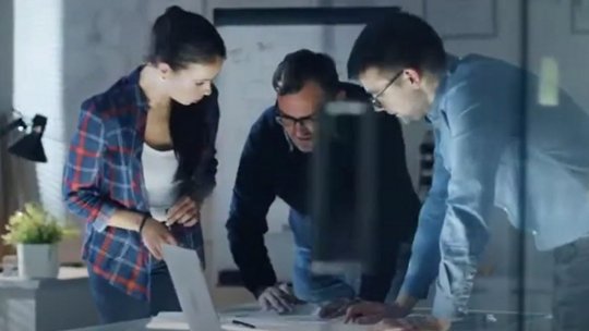 Three people in business casual attire gathered around a laptop, reviewing documents at a table in a modern office.