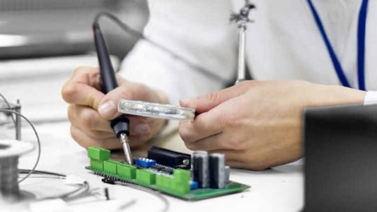 A person is soldering components on a green circuit board in a well-lit workspace