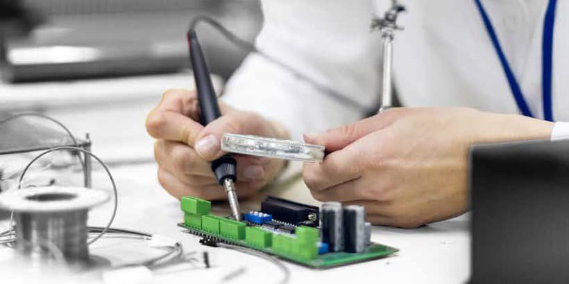A person is soldering components on a green circuit board in a well-lit workspace